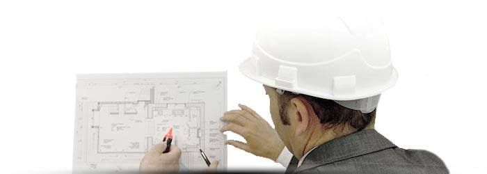 Un ouvrier portant un casque blanc examine un plan d'étage et montre des plans sur un mur.