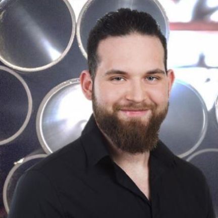 Ein Mann mit Bart trägt ein schwarzes Hemd und lächelt vor einer Wand aus Rohren.