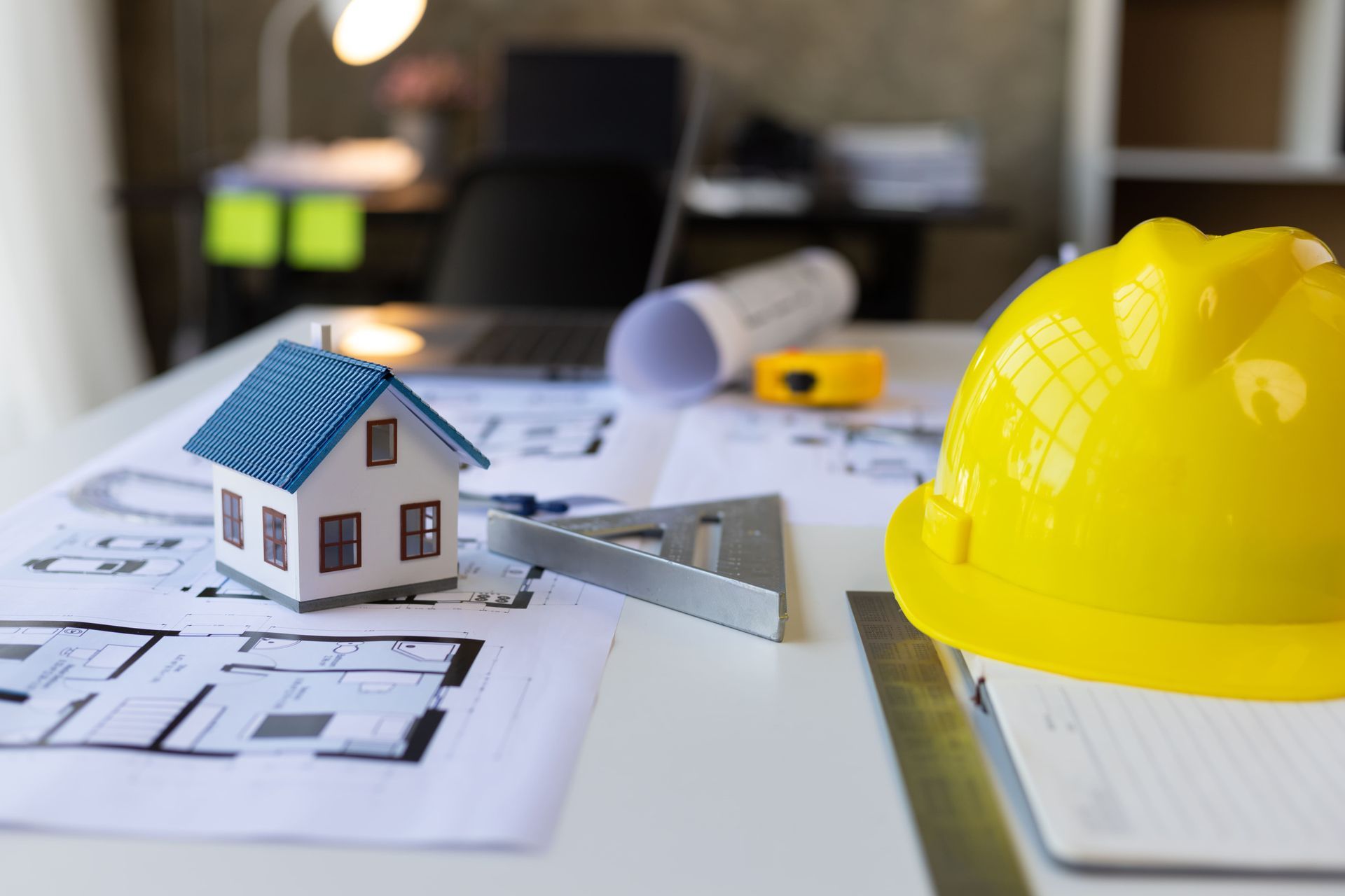 Blueprints with a small house model, ruler, and yellow hard hat on a desk.