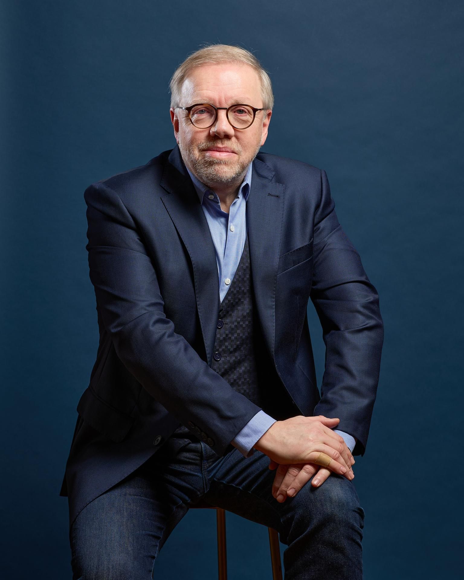 Homme en blazer et gilet, assis sur un tabouret, regardant le spectateur. Décor bleu marine et denim.