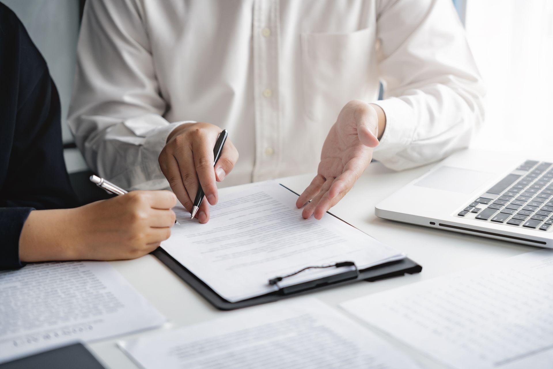 Deux personnes examinent des documents, l'une d'elles signe avec un stylo, un ordinateur portable est visible en arrière-plan.