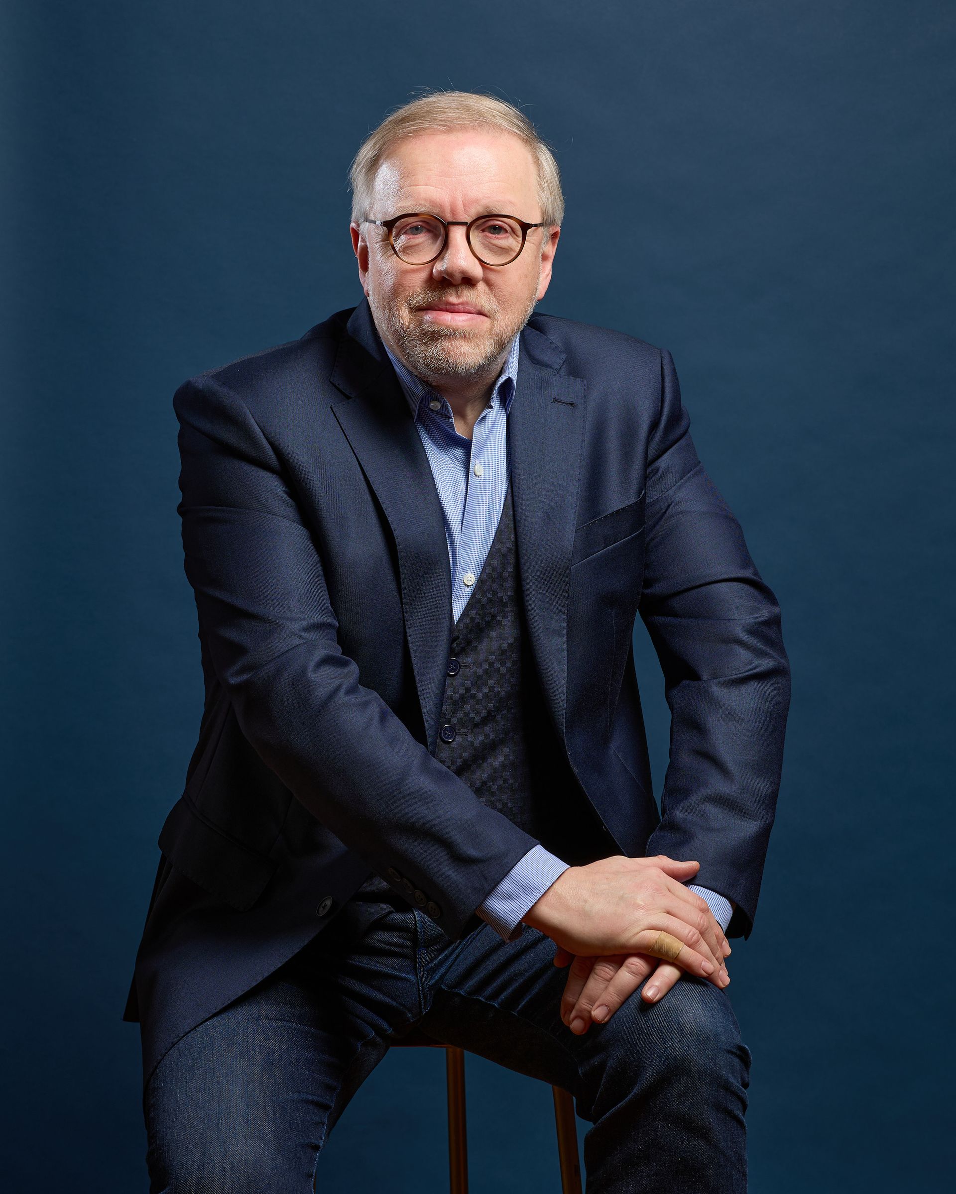 Homme assis, vêtu d'un blazer bleu, d'un gilet, d'une chemise et d'un jean, regardant le spectateur. Fond bleu foncé.