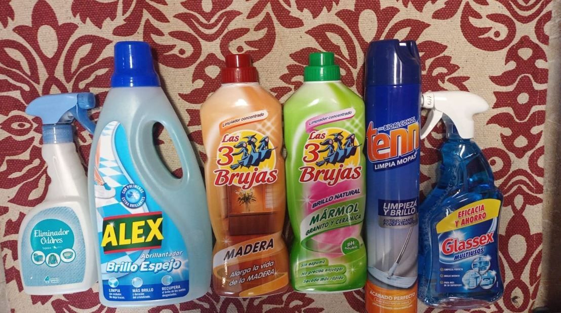 Artículos de limpieza dispuestos sobre una superficie estampada, con botellas y pulverizadores de varias marcas y colores.