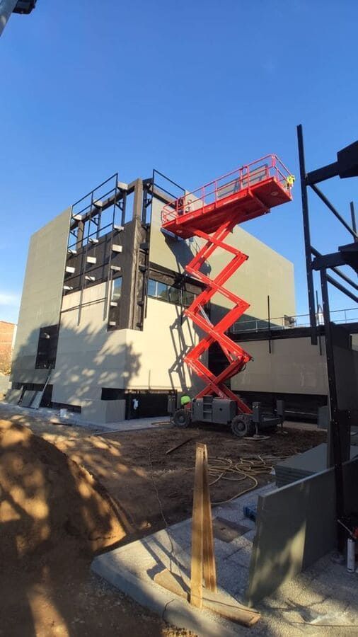 Elevador de tijera rojo junto a un edificio en construcción; cielo azul.