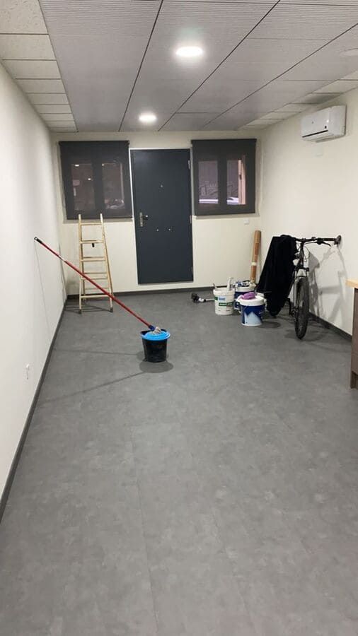 Habitación vacía con suelo gris, paredes blancas, dos ventanas, puerta y artículos de limpieza. También hay una bicicleta.