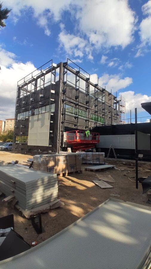 Sitio de construcción: edificio con estructura de acero en construcción, trabajadores en el ascensor, materiales en el suelo.