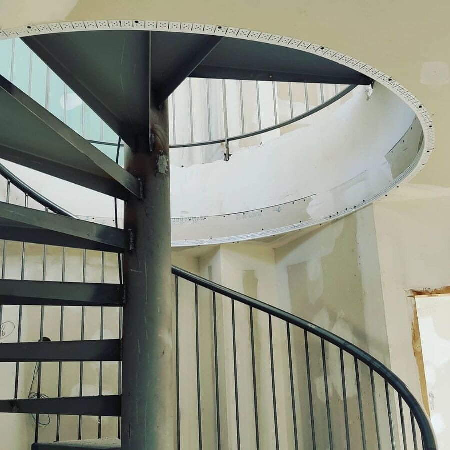 Escalera de caracol con barandilla metálica y soporte central. Paredes blancas al fondo.