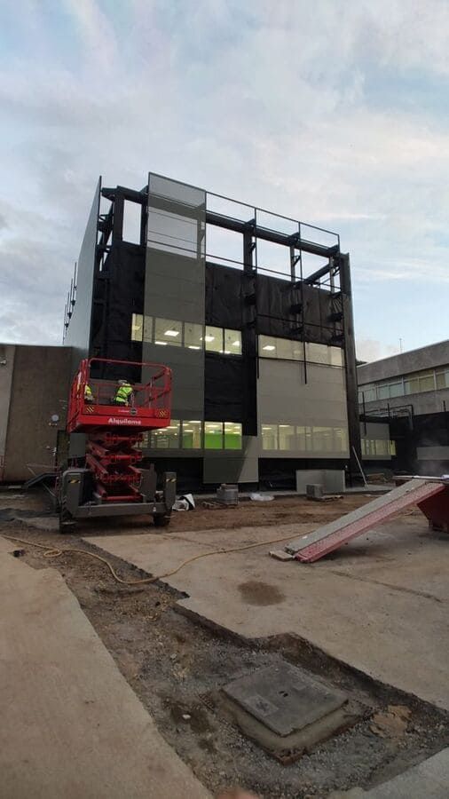 Obra con un edificio de varias plantas a medio construir; trabajador en un ascensor. Paneles grises y negros, cielo nublado.
