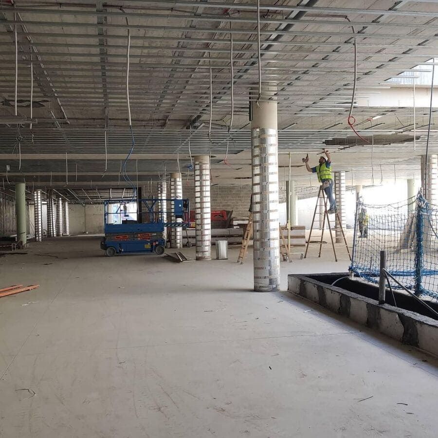 Interior de una obra en construcción, techo sin terminar, trabajadores, andamios y ascensor. Suelo de hormigón y estructura metálica.