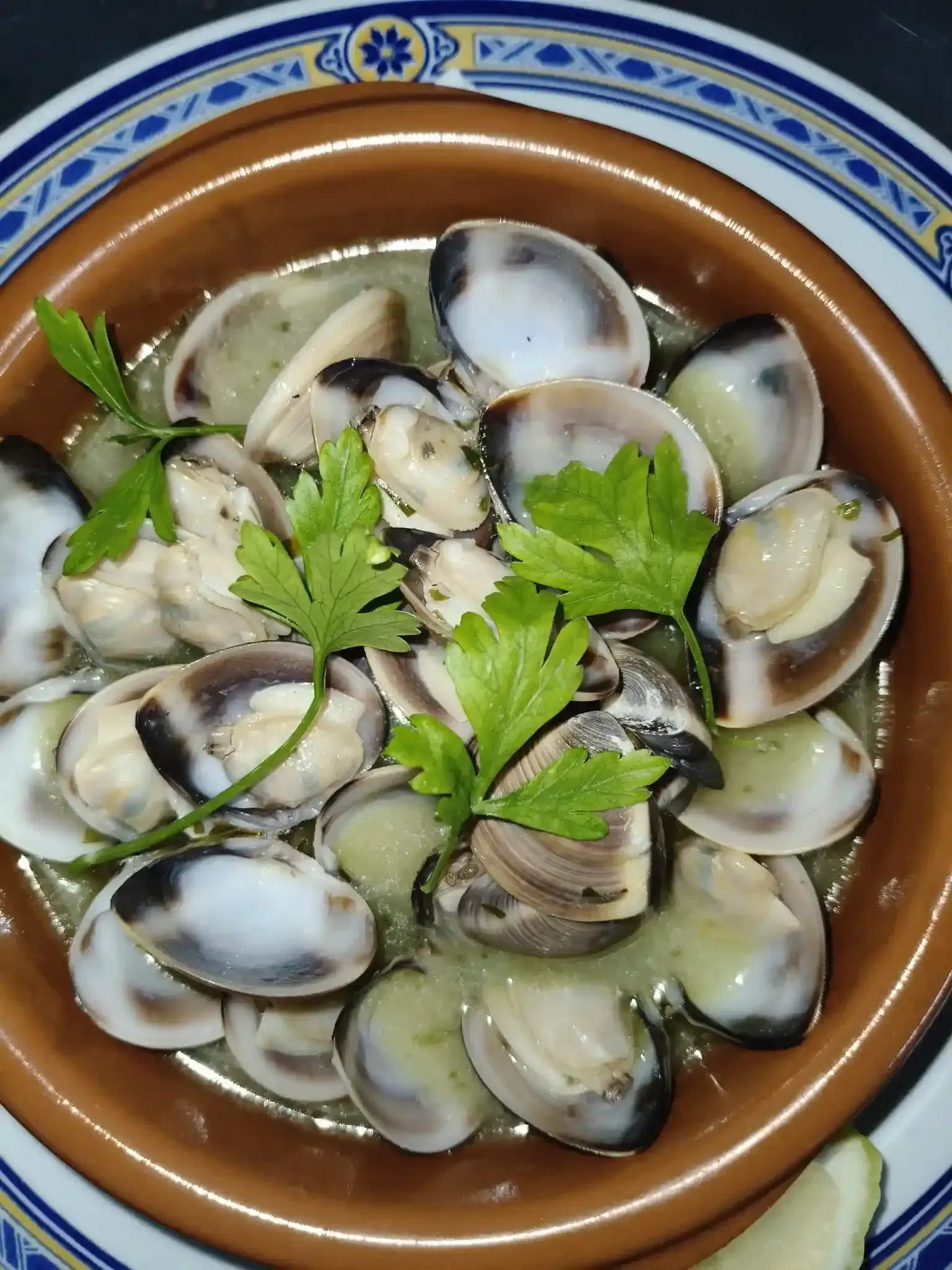 Almejas en un recipiente de cerámica marrón, adornadas con perejil, servidas en una salsa ligera, en un plato.