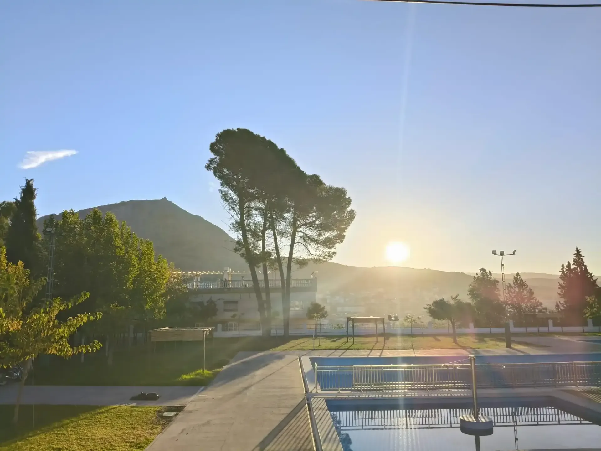Vista de la piscina al amanecer con montañas al fondo, algunos árboles y edificios.