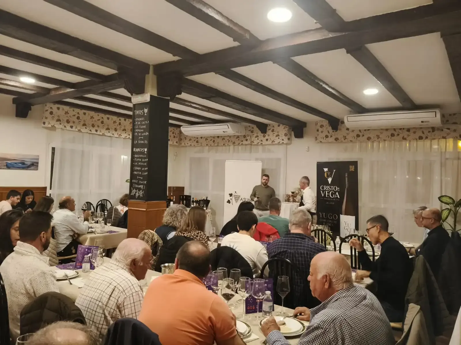 La gente sentada en las mesas escucha una presentación en un restaurante, con los presentadores de pie cerca de una pancarta.