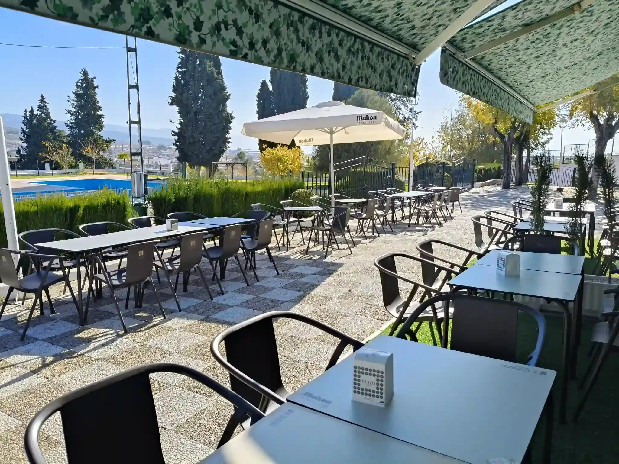 Cafetería al aire libre con mesas y sillas bajo toldos, con vistas a la piscina y a los árboles.
