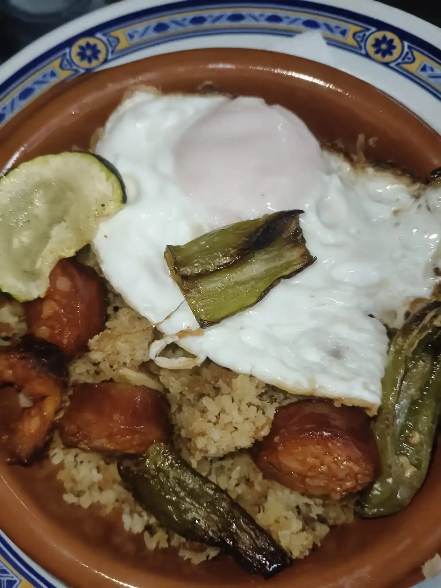 Plato con arroz, salchicha, pimientos, calabacín y huevo frito. Plato marrón, huevo blanco, comida.