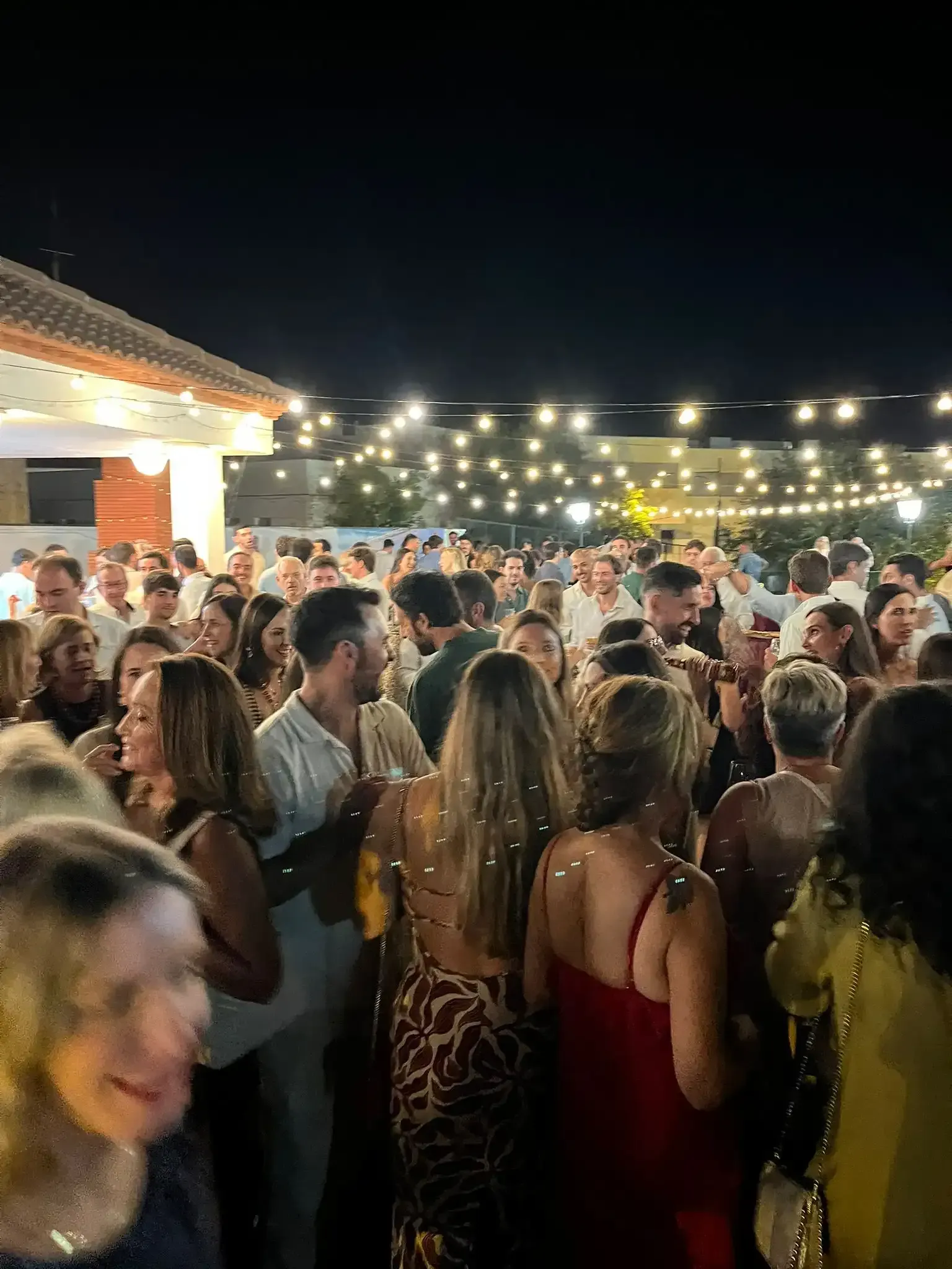 Fiesta nocturna con mucha gente, ambiente al aire libre, luces de cadena, ambiente festivo.