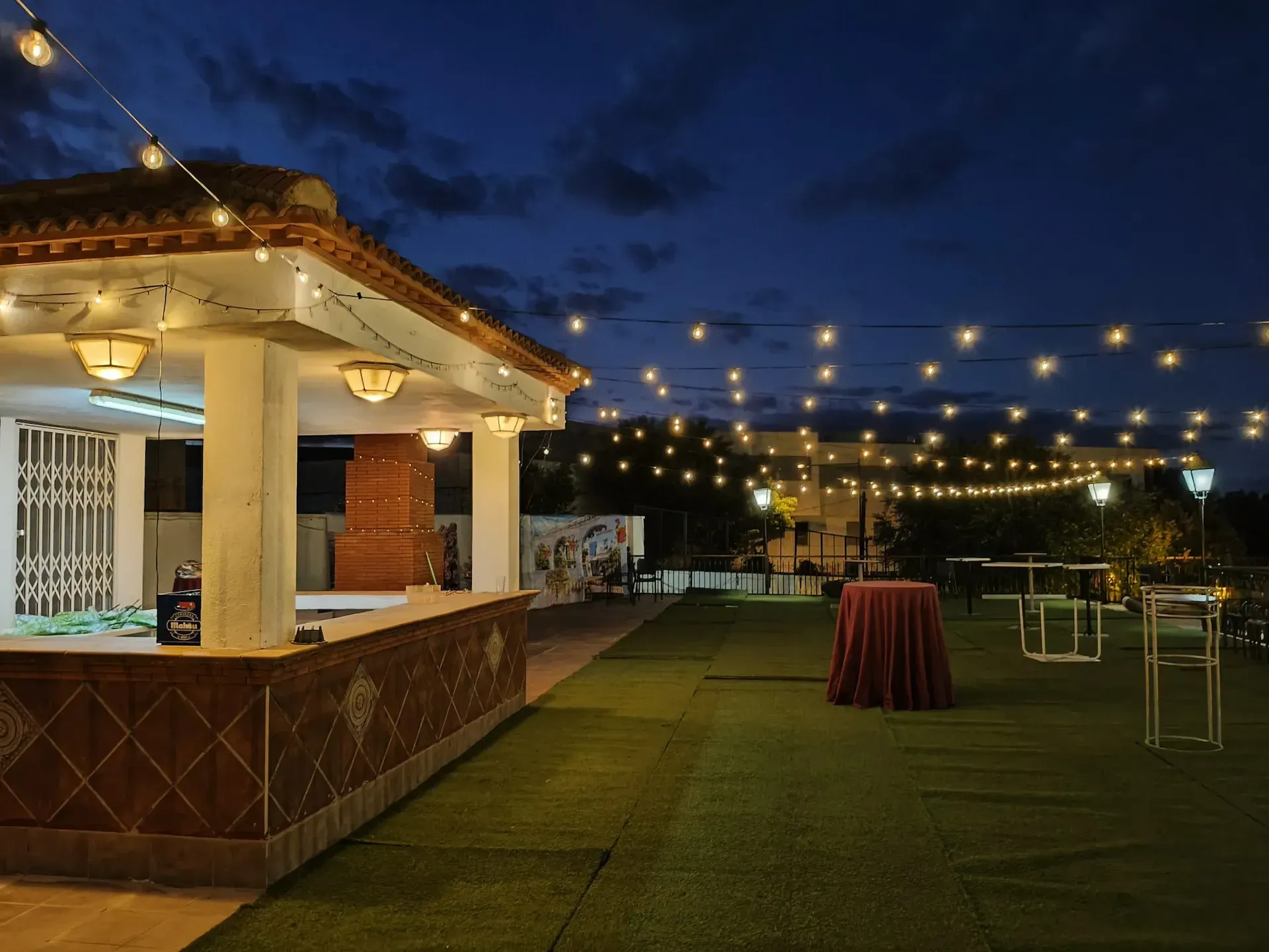 Cafetería al aire libre con mesas y sillas bajo toldos, con vistas a la piscina y a los árboles.