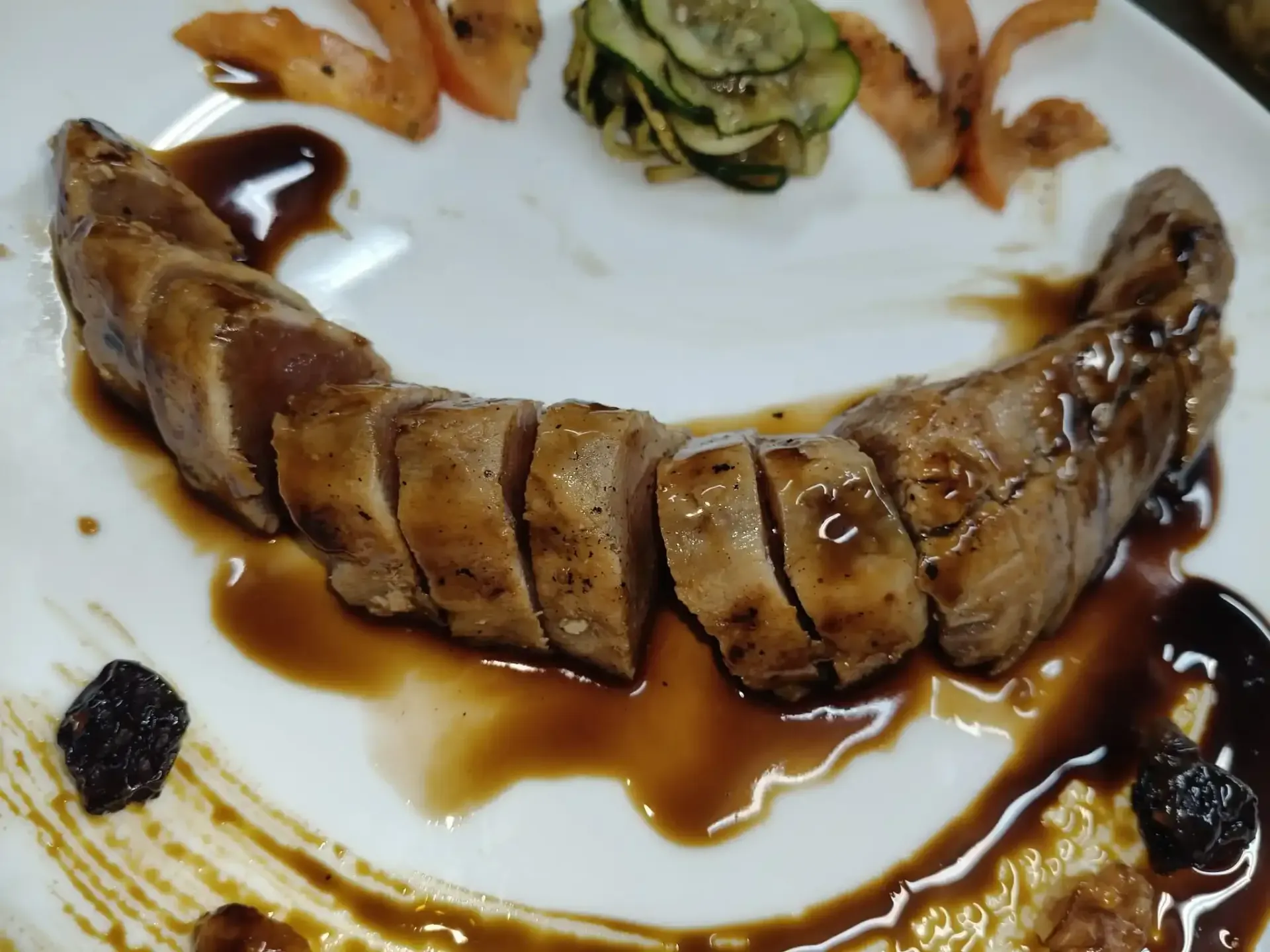 Solomillo de cerdo en rodajas con salsa, tomates y pepinos en un plato blanco.