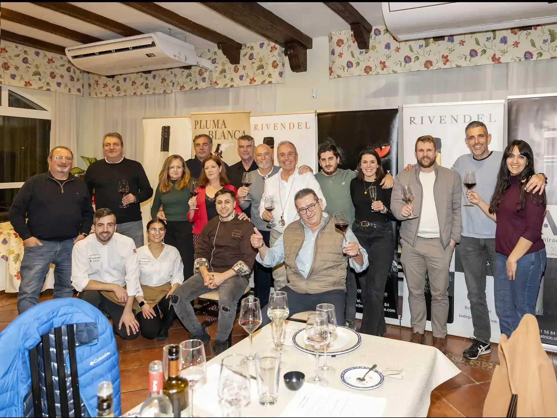 Un grupo de personas, incluyendo personal, posa en un restaurante. Algunos sostienen copas de vino, sonriendo.