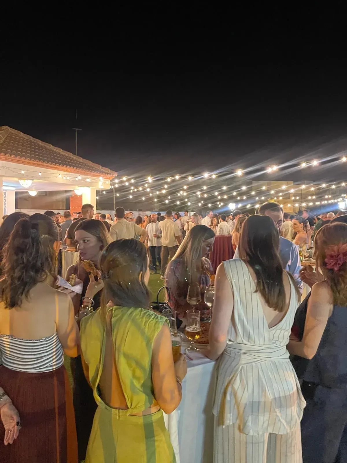 Evento nocturno: Los invitados se mezclan bajo luces de colores, cerca de un edificio.