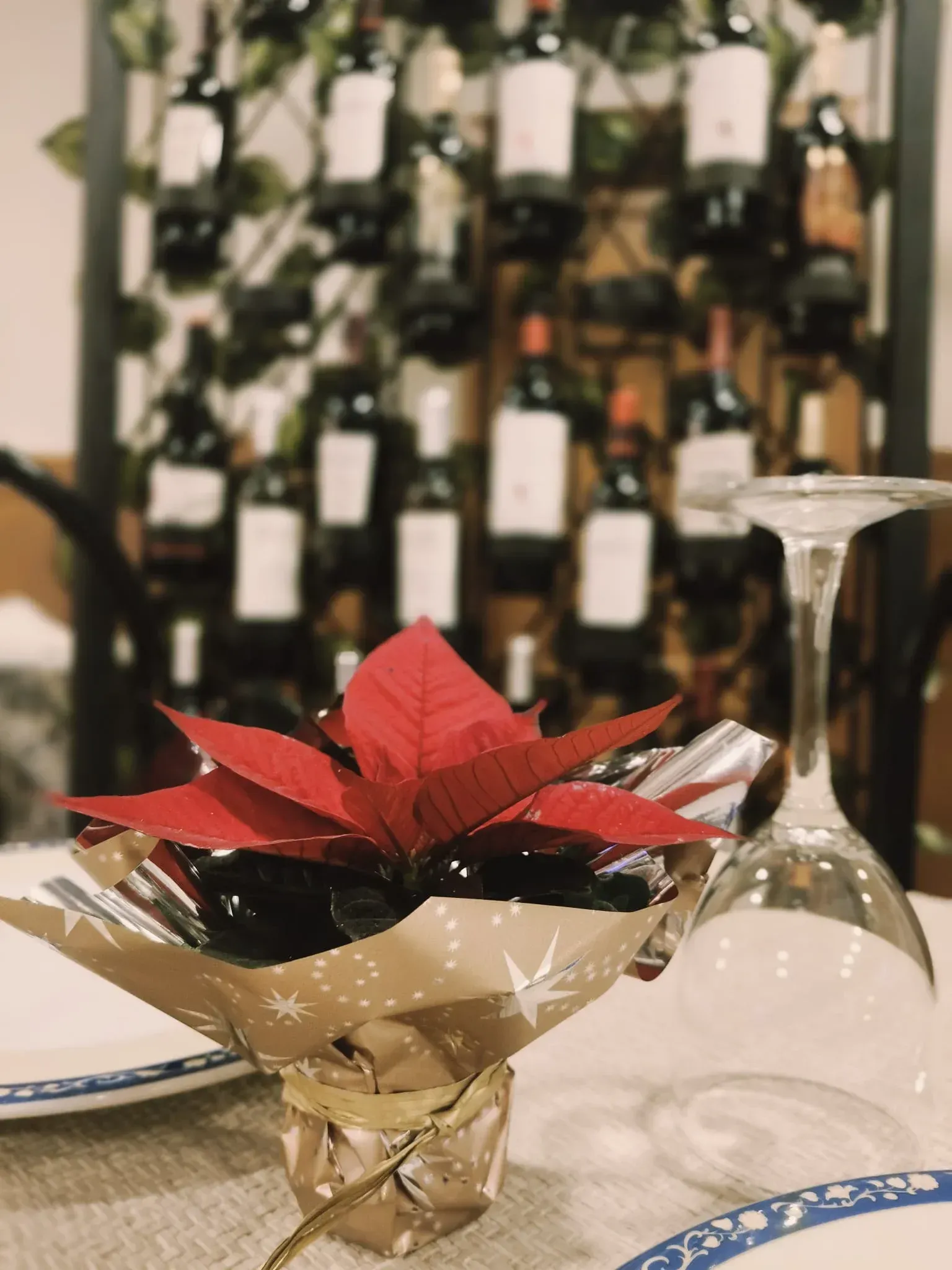 Centro de mesa con flor de Pascua roja, botellas de vino al fondo. Copas y platos presentes.