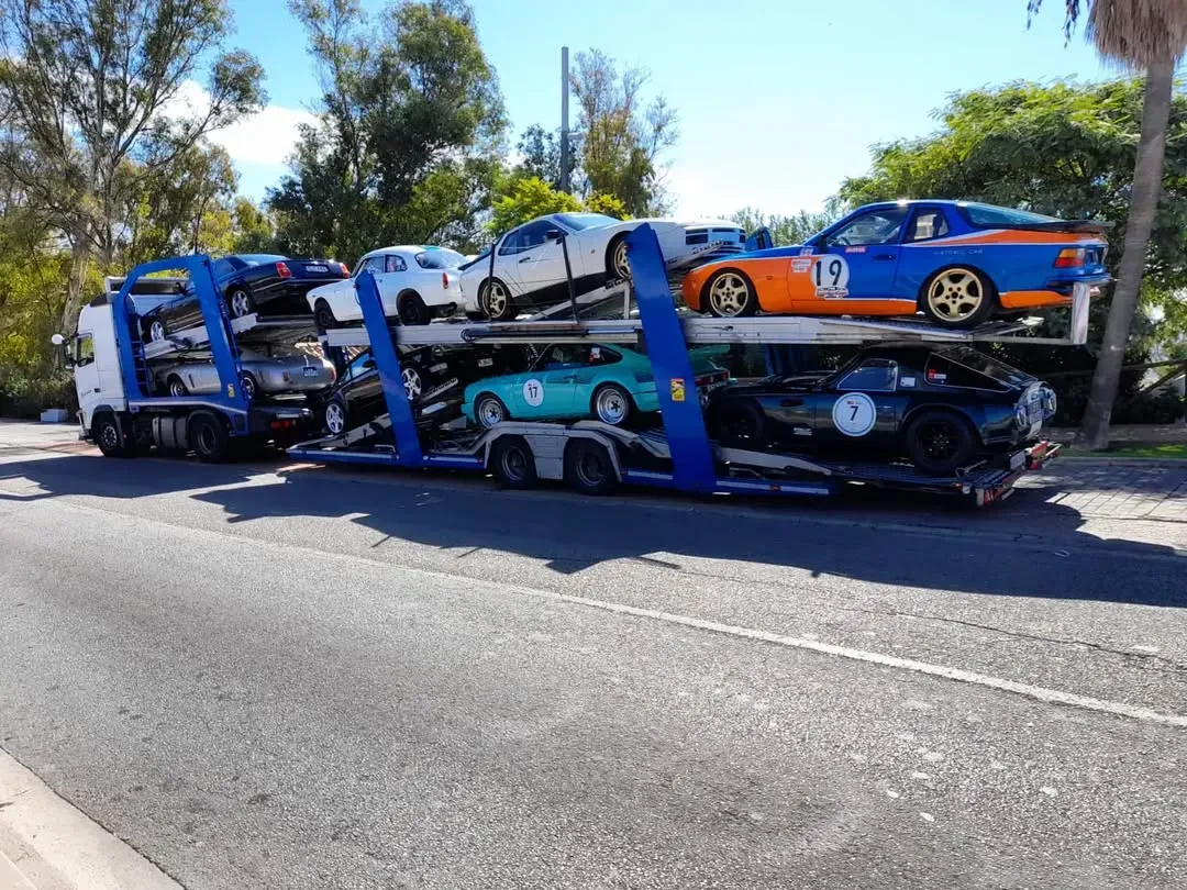 Camión transportador de automóviles cargado con varios coches deportivos antiguos en una carretera durante el día.