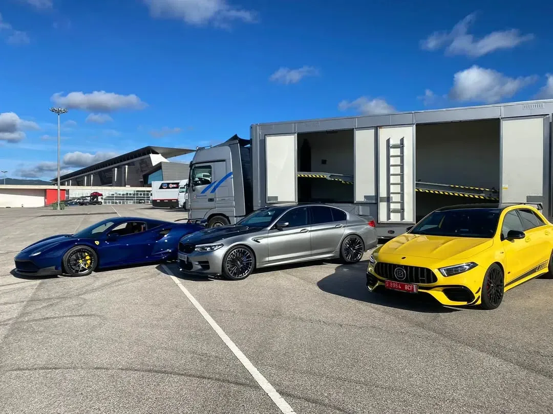 Tres coches de lujo (un Ferrari azul, un BMW plateado y un Mercedes amarillo) aparcados cerca de un camión semirremolque con portacoches.