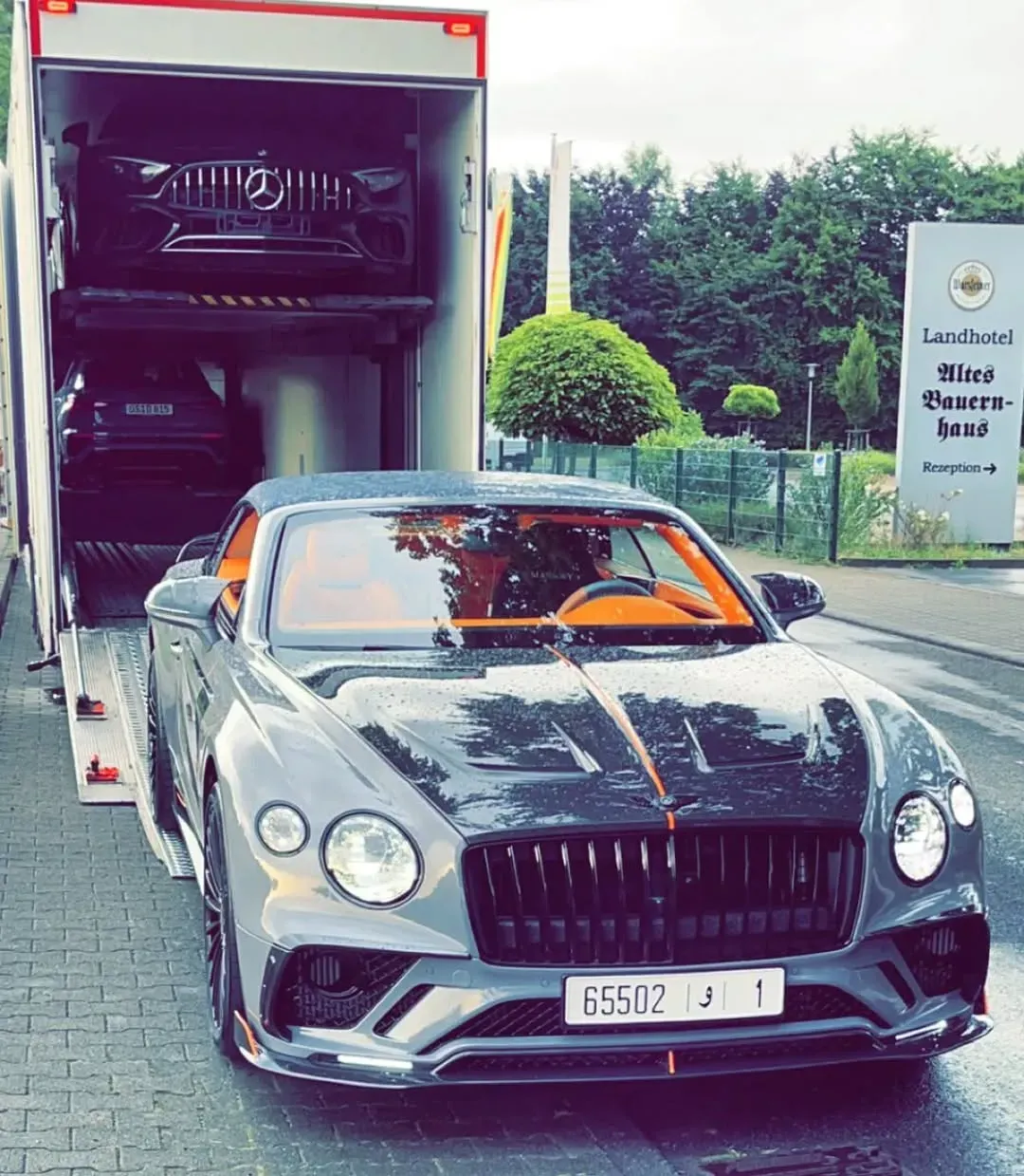 Bentley convertible con interior naranja, estacionado delante de un camión de transporte cargado de coches de lujo.