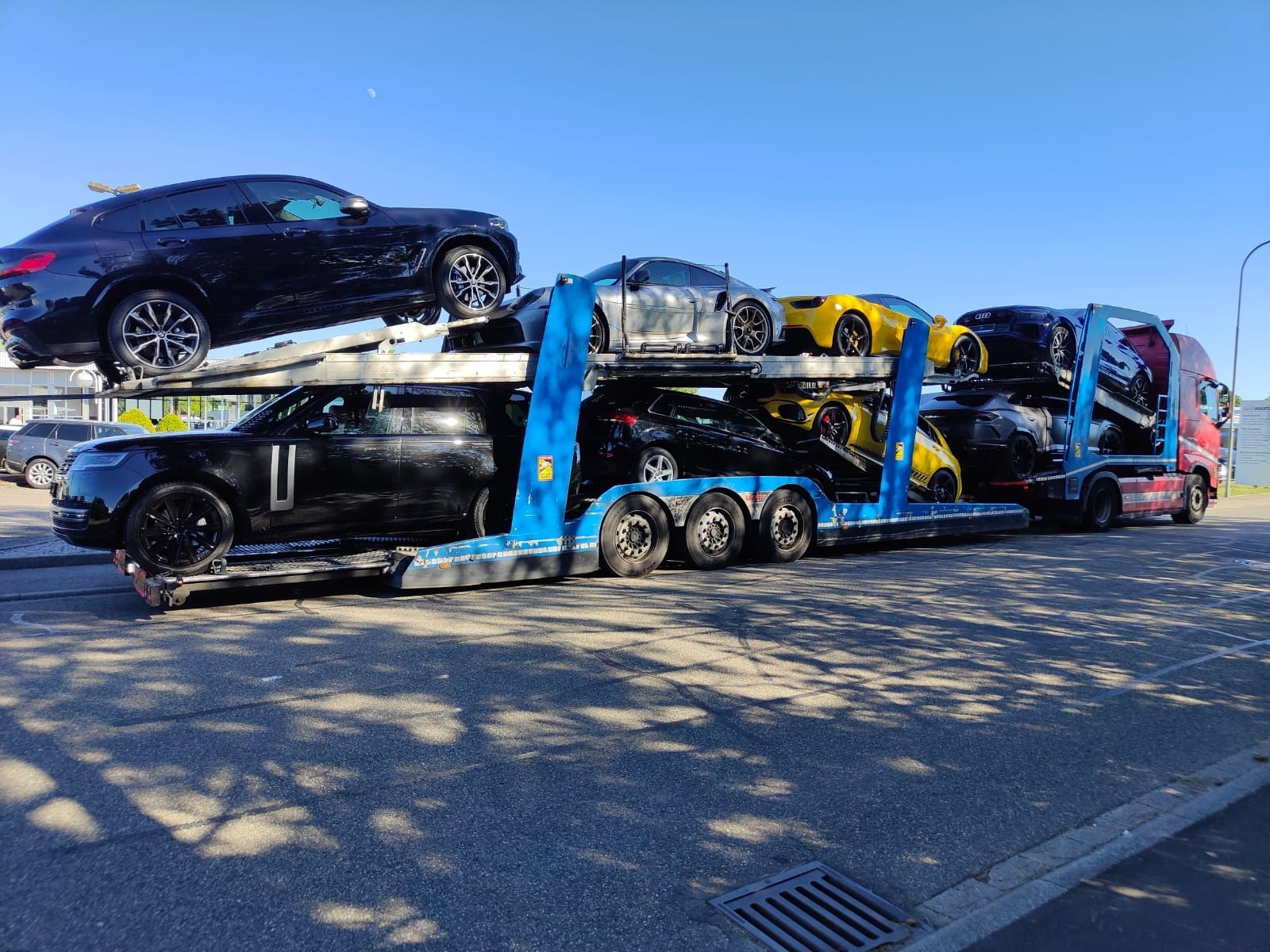 Un camión transportador de automóviles cargado con varios vehículos de lujo en varios colores, estacionado al aire libre en un día soleado.