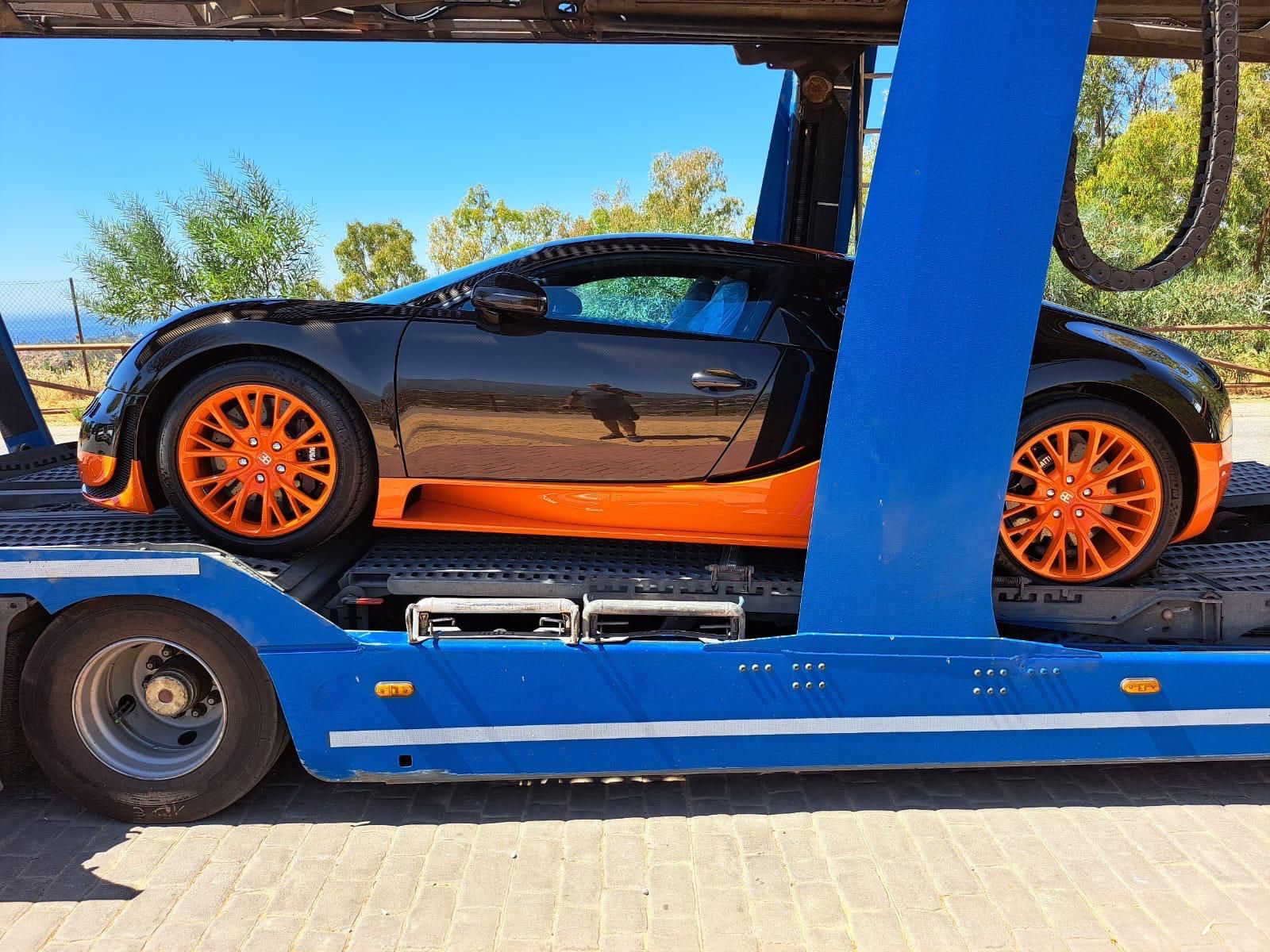 Bugatti Veyron negro y naranja en un transportador de automóviles azul, al aire libre.