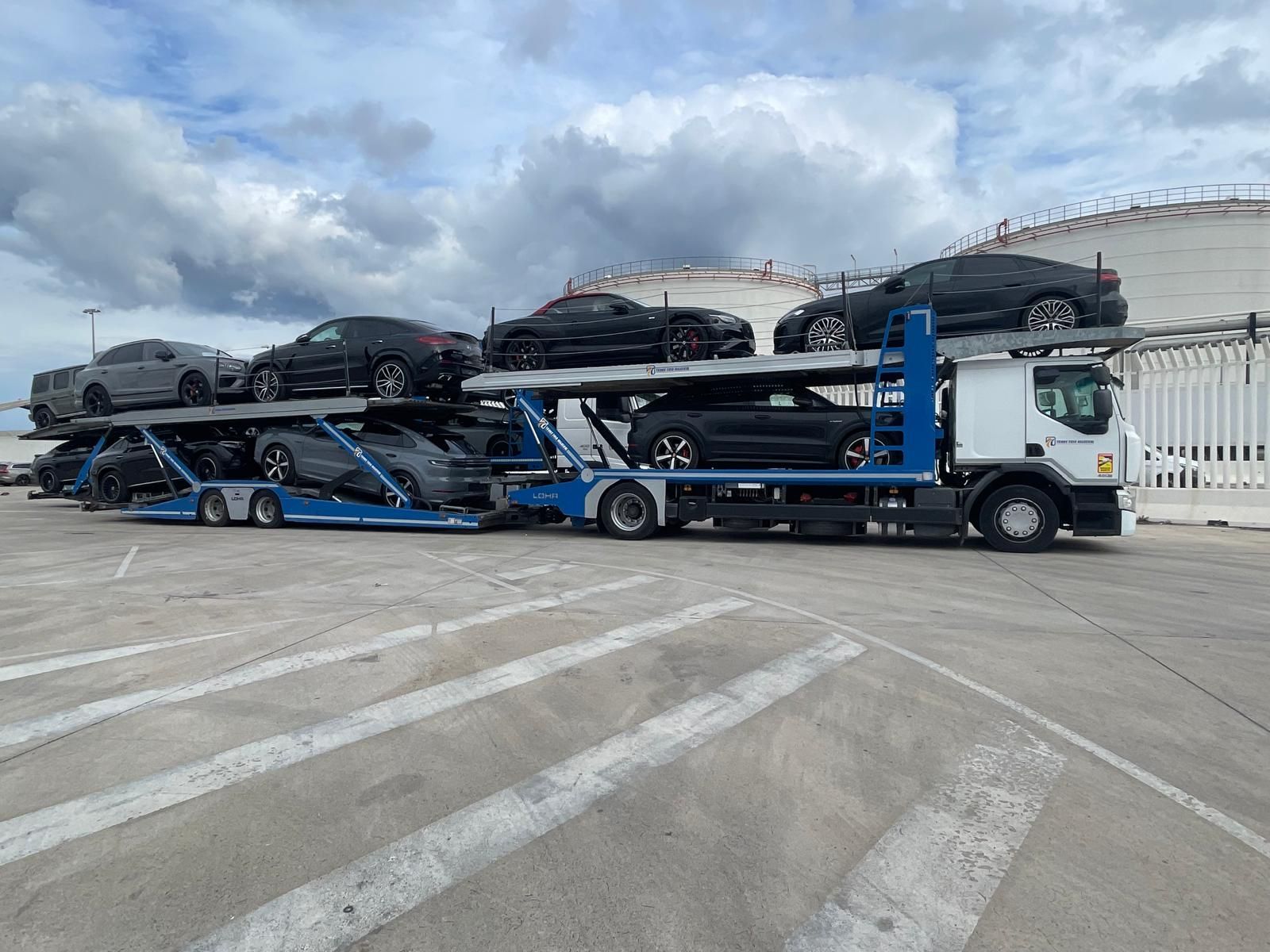 Camión transportador de automóviles cargado con vehículos negros estacionados sobre concreto, cielo nublado.