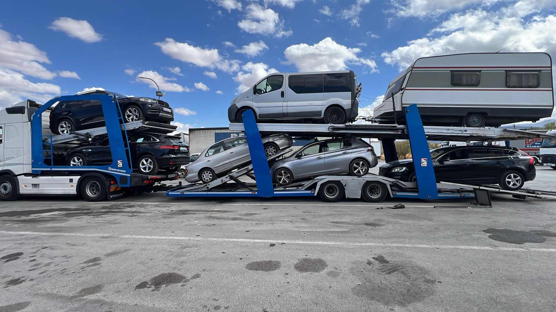 Camión que transporta varios automóviles, una camioneta y una caravana en un día soleado en un estacionamiento.
