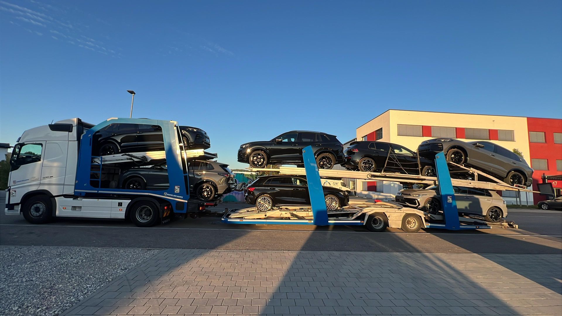 Camión que transporta varios todoterrenos negros en un día soleado, estacionado cerca de un edificio con detalles en rojo.