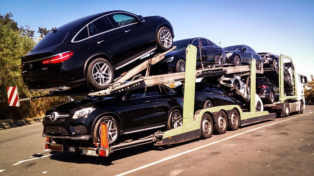 Un camión transportador de automóviles transporta varios vehículos Mercedes-Benz negros por una carretera, bajo un cielo despejado.