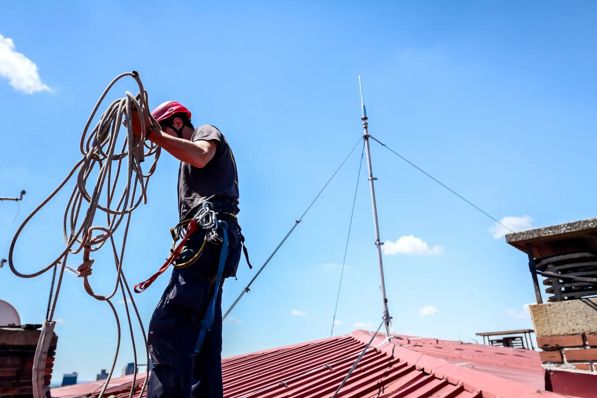 Techador con arnés de seguridad sobre un techo rojo, sosteniendo cuerdas cerca de un pararrayos bajo un cielo azul.