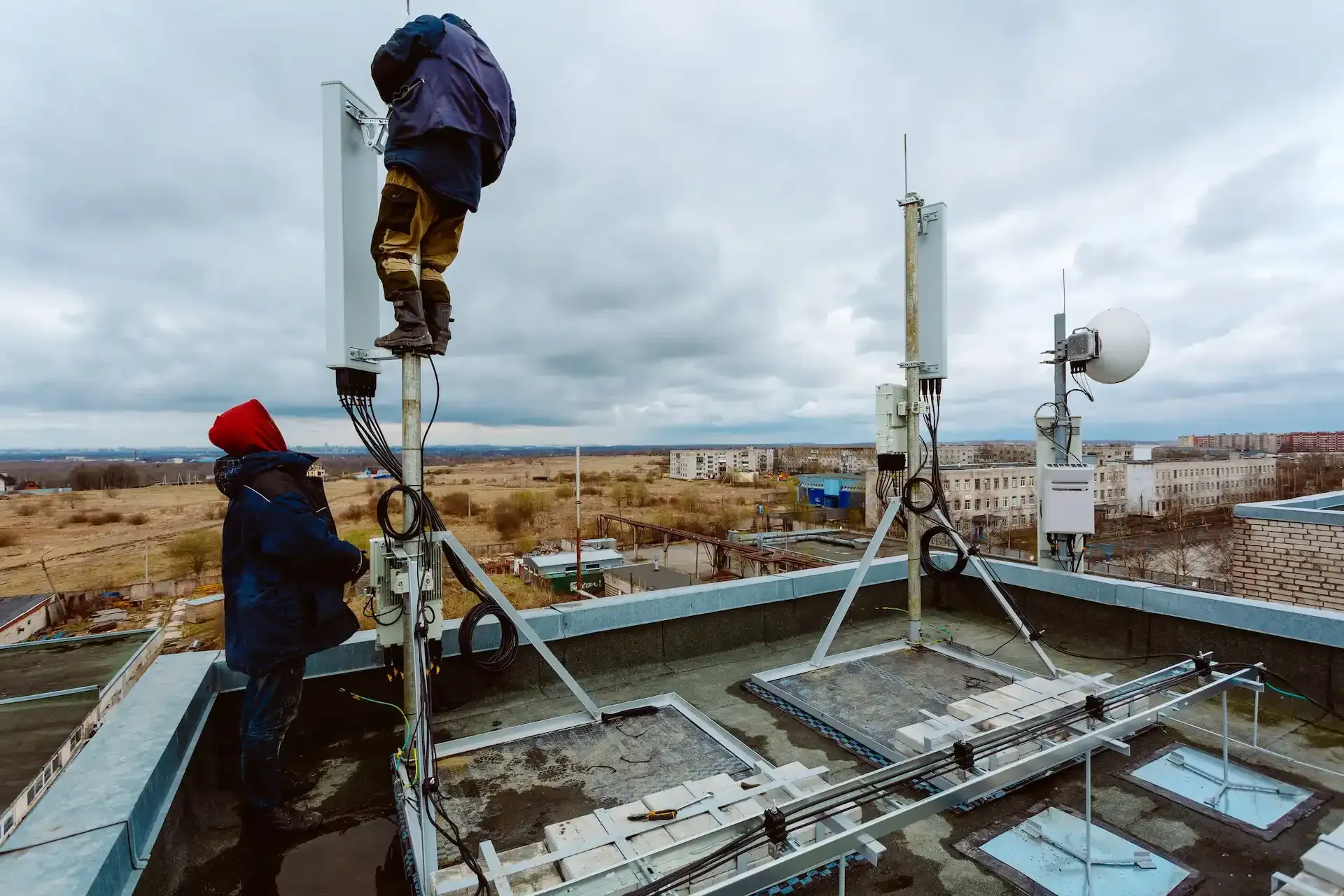 Dos técnicos trabajan en una antena celular en un tejado con un cielo nublado de fondo.