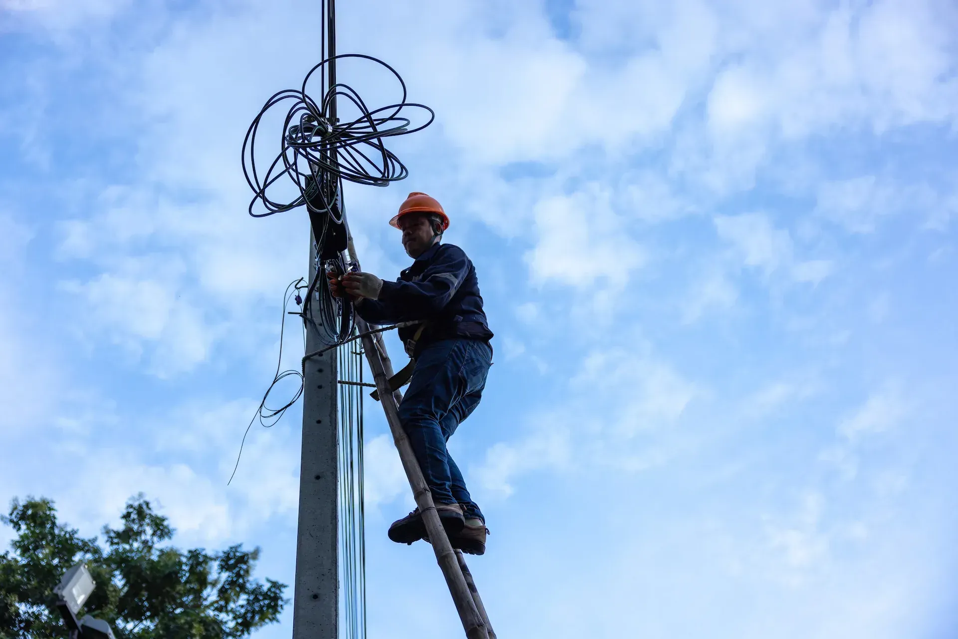 Un liniero con casco naranja trabaja con cables encima de un poste de servicios públicos contra un cielo nublado.