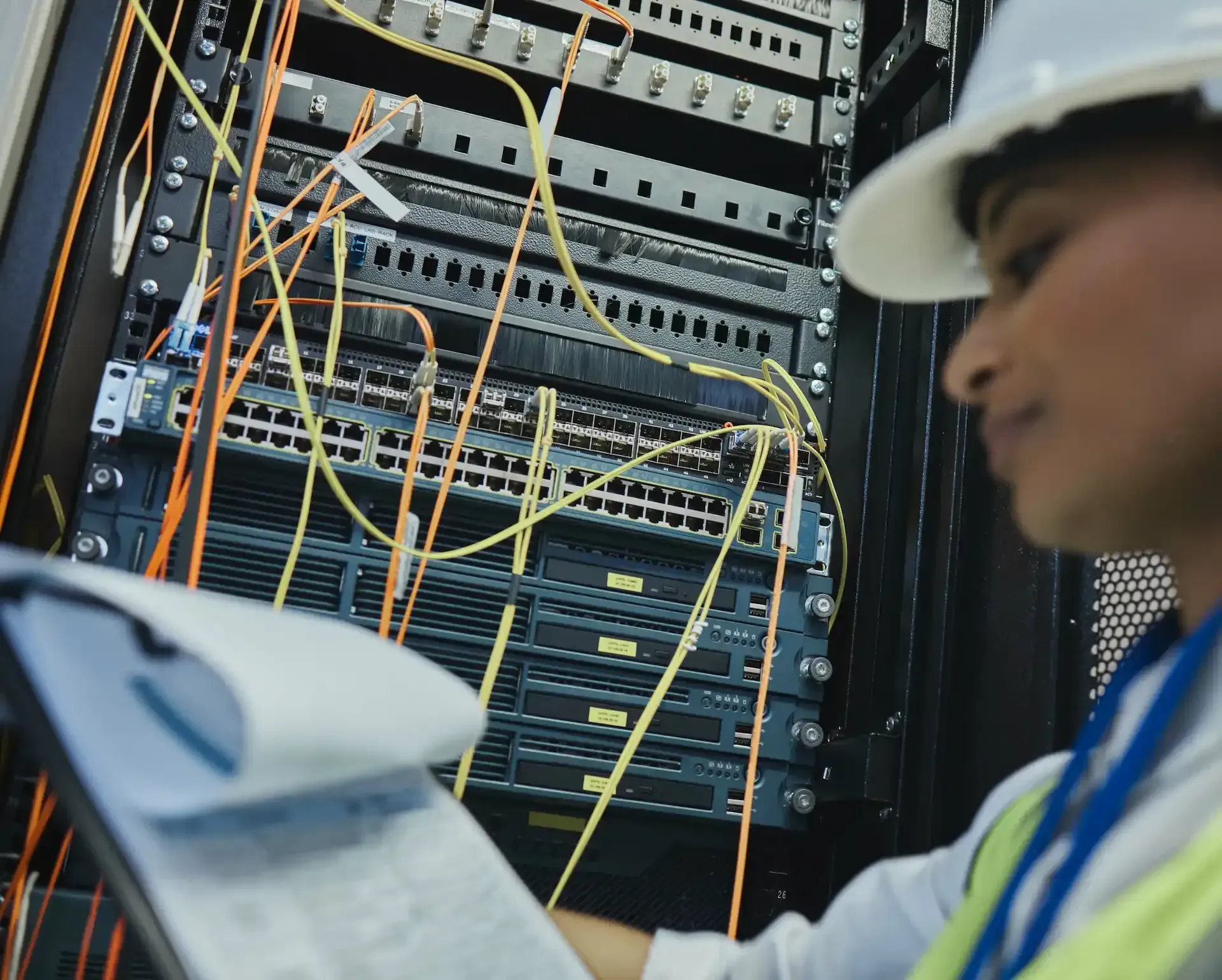 Una persona con casco examina el rack del servidor y toma notas. Los cables están conectados al equipo.