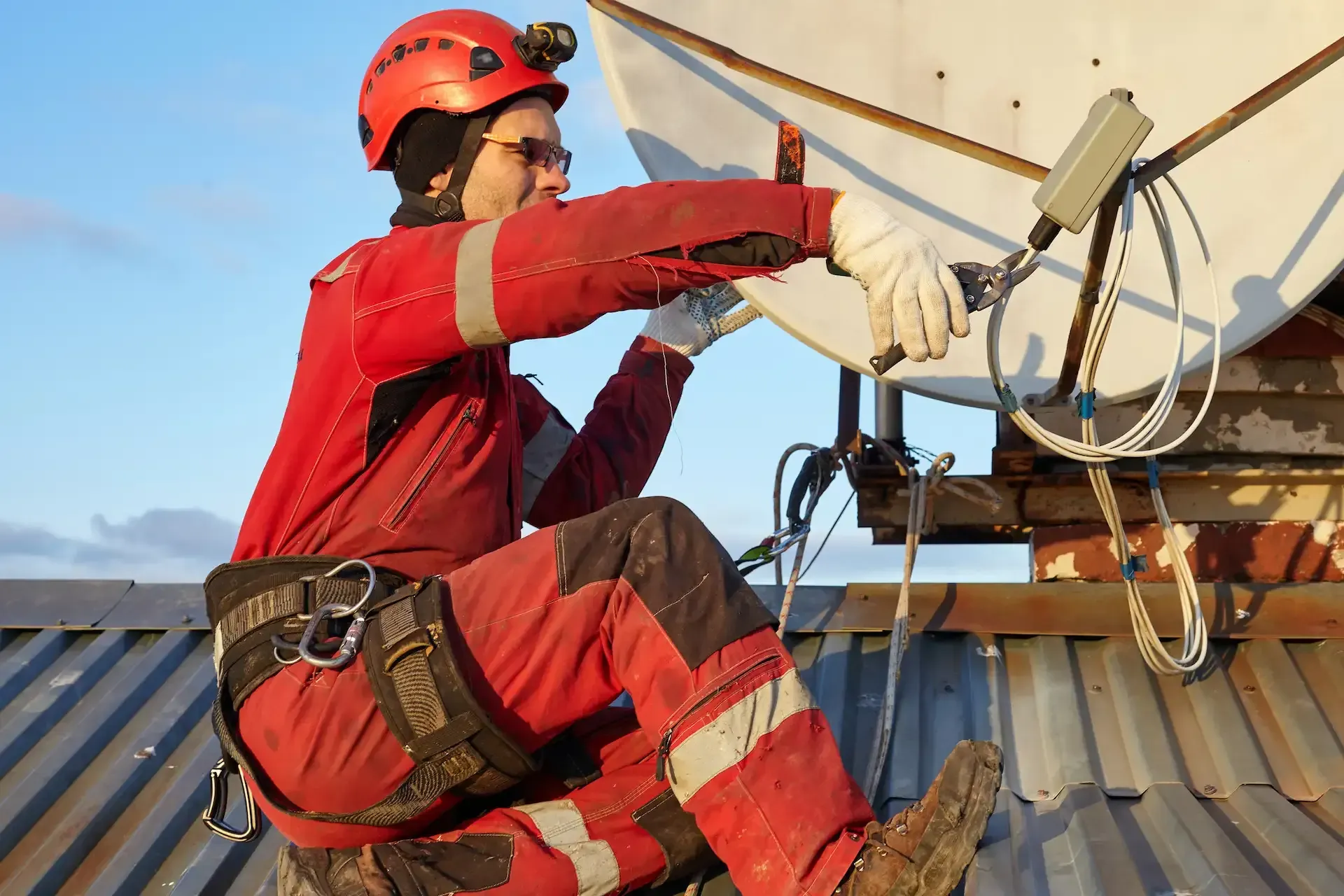 Persona con overol rojo en un tejado instalando una antena parabólica. Lleva casco, guantes y arnés de seguridad.