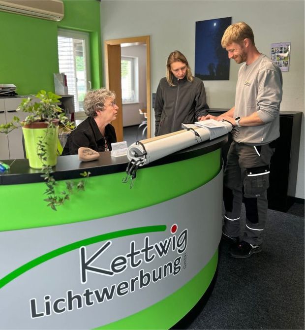 Eine Frau sitzt an einem Schreibtisch vor einem Schild mit der Aufschrift „Kettwig Lichtwerbung“