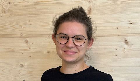 Eine Frau mit Brille und schwarzem Hemd steht vor einer Holzwand.