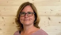 Eine Frau mit Brille und rosa Hemd steht vor einer Holzwand.