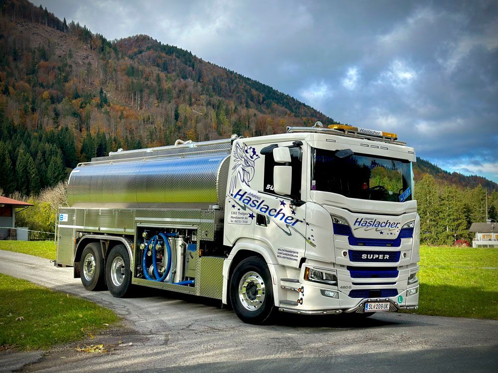Ein weißer Scania-Tankwagen mit der Aufschrift „Haslacher“ parkt auf einer Straße vor der Kulisse bewaldeter Berge.