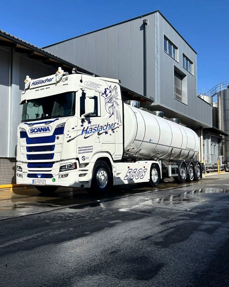 Ein weißer Scania-Sattelzug mit großem Tankauflieger parkt vor einem modernen Industriegebäude unter blauem Himmel.