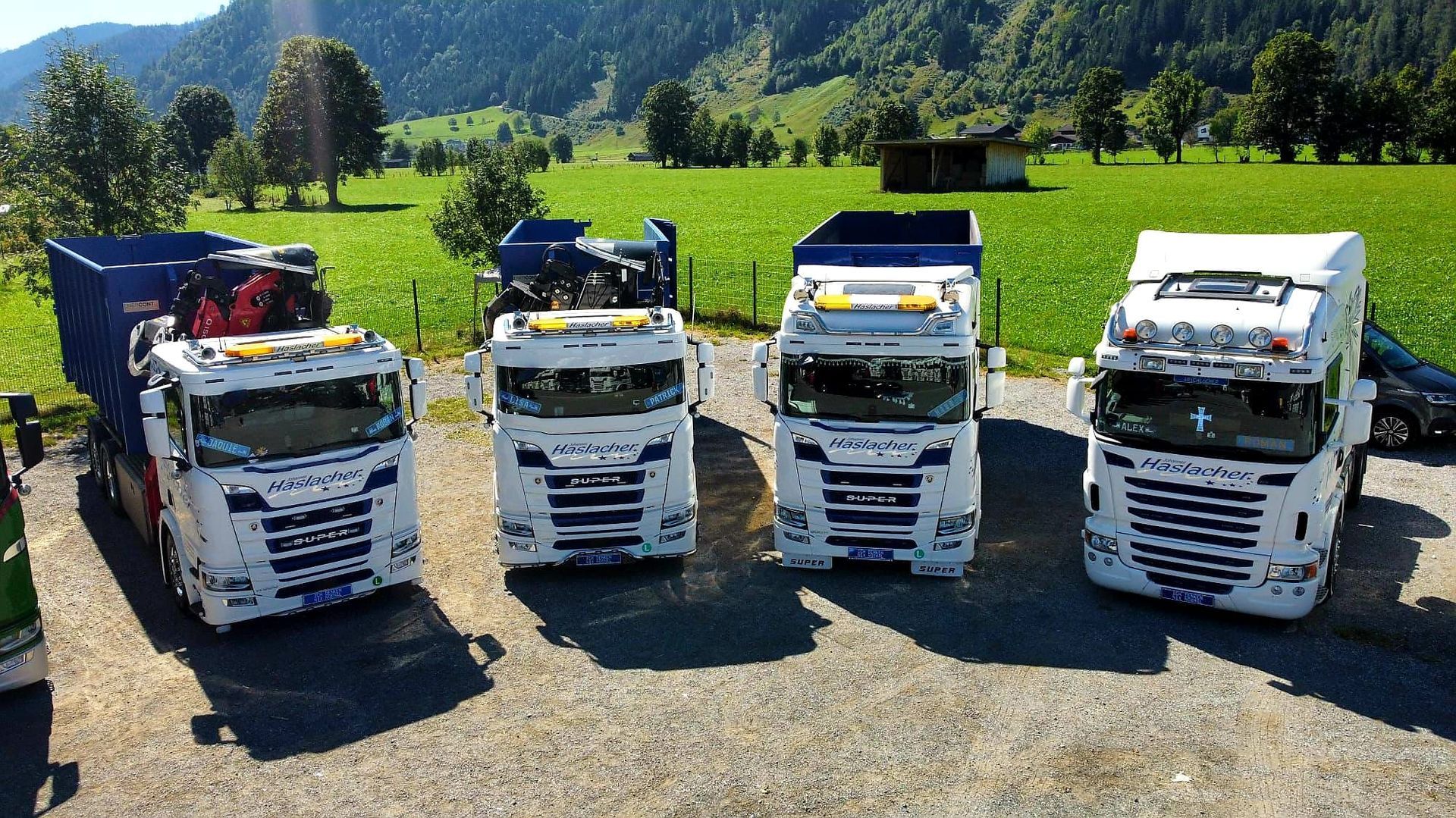 Vier weiß-blaue Sattelzüge parken auf dem Gehweg, im Hintergrund grüne Felder und Berge.