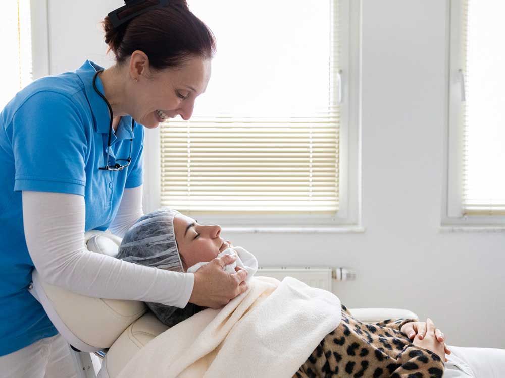 Eine Frau im blauen Hemd führt eine Gesichtsbehandlung bei einer Frau die auf der Behandlungsliege liegt durch