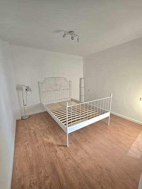 Habitación vacía con estructura de cama de metal blanco y suelo de madera, iluminada por una lámpara de techo.