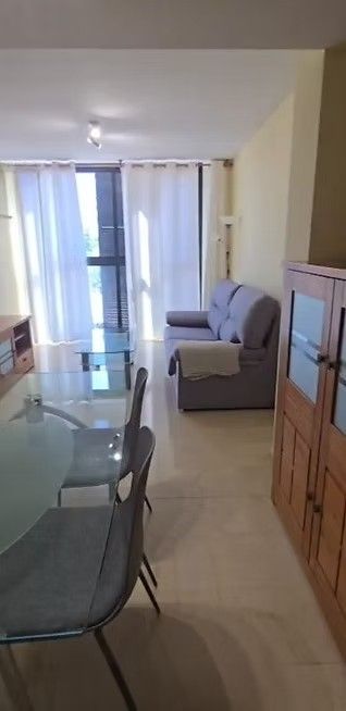 Luminoso salón con mesa de comedor de cristal, sofá, armario de madera y cortinas transparentes junto a la puerta del balcón.