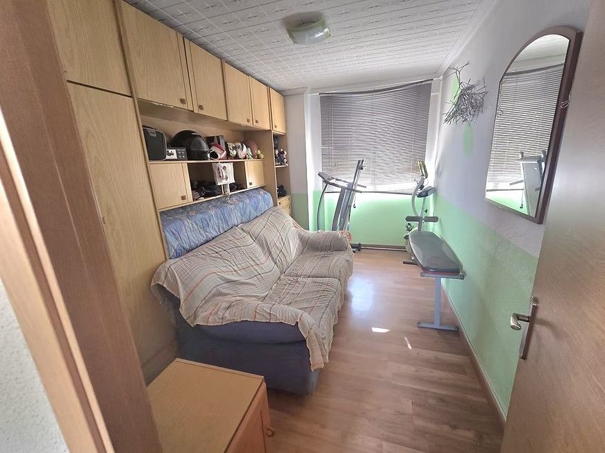 Habitación estrecha con sofá cama, armarios de madera, estantes y una ventana con persianas y cortina de rayas.