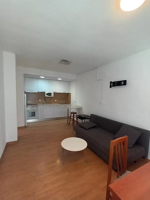 Pequeña sala de estar con sofá gris, mesa de centro redonda blanca, suelo de madera y cocina compacta al fondo.