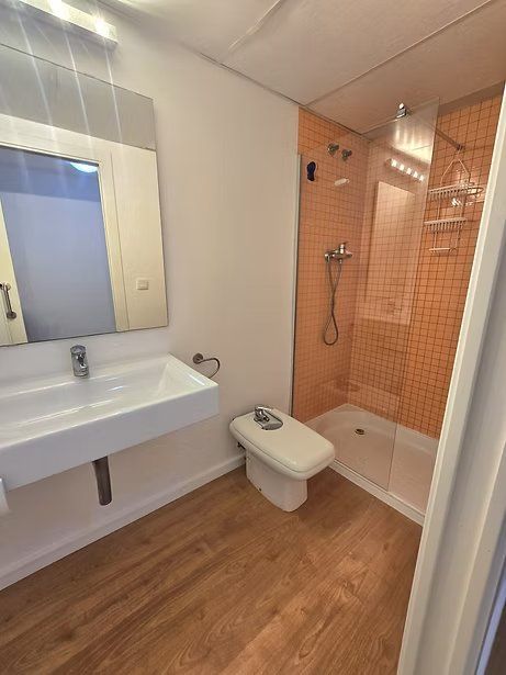 Baño pequeño con lavabo blanco, inodoro, suelo de imitación madera y cabina de ducha con azulejos naranjas.